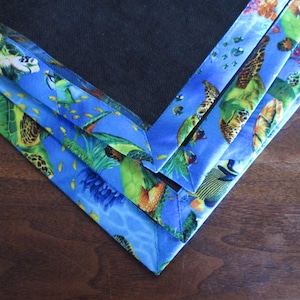 May include: Close-up of a blue fabric with a sea turtle and aquatic life pattern. The fabric is folded in a V-shape, revealing a dark mesh material. The edges are trimmed with a vibrant blue border featuring sea turtles, fish, and coral.