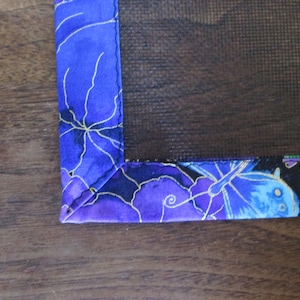 May include: Close-up of a corner of a decorative fabric item. The fabric features a vibrant blue background with a floral pattern in shades of purple and gold. The edges are neatly sewn, showcasing the item's craftsmanship.