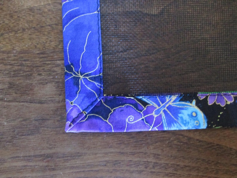 May include: Close-up of a corner of a decorative fabric item. The fabric features a vibrant blue background with a floral pattern in shades of purple and gold. The edges are neatly sewn, showcasing the item's craftsmanship.