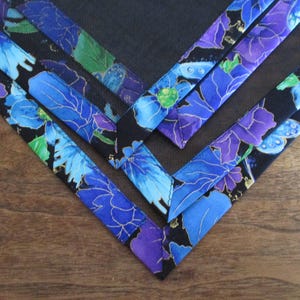 May include: Close-up of several fabric corners with a floral pattern. The fabric features blue and purple flowers with gold accents on a black background. The corners are arranged in a V-shape on a wooden surface.
