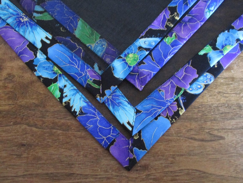 May include: Close-up of several fabric corners with a floral pattern. The fabric features blue and purple flowers with gold accents on a black background. The corners are arranged in a V-shape on a wooden surface.