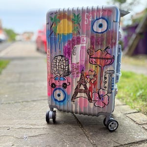 May include: A silver travel suitcase decorated with vibrant graffiti art. The design features a sunset, palm trees, an eye symbol, a car, the Eiffel Tower, and a cocktail. The case has four wheels and a retractable handle.