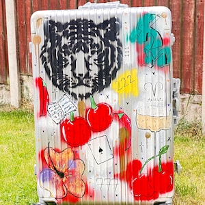 May include: Silver rolling suitcase with graffiti art. Features a tiger face, cherries, a playing card, and the numbers 222 and 777. The suitcase is on a grassy surface.