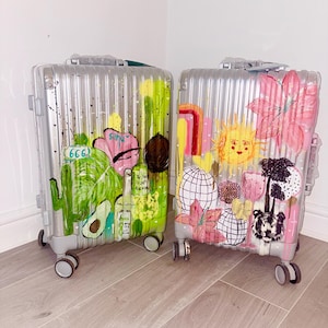 May include: Two silver hard-shell suitcases with vertical ridges and four wheels. The left suitcase features green painted designs including an avocado, a cactus, and the number 666. The right suitcase features pink and yellow painted designs including a sun, flowers, a tiger face, and disco balls.