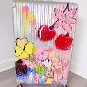 May include: A silver hard-shell suitcase featuring a vibrant hand-painted design. The artwork includes large red cherries, a yellow hibiscus flower, a palm tree, a sailboat, the Eiffel Tower, and two cocktail glasses with the word Summer printed.