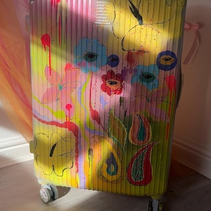 Op de afbeelding: Een harde koffer met een verticaal geribbelde textuur, voorzien van een handgeschilderd ontwerp van kleurrijke bloemen, abstracte vormen en oogmotieven in tinten geel, roze, blauw en groen met zichtbare verfdruppels.