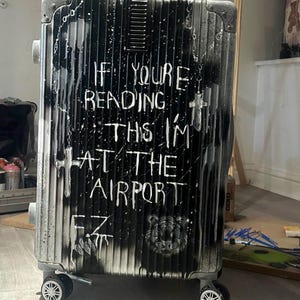 May include: A hard-shell suitcase with a vertical ribbed texture, featuring a black and white spray-painted design. The text reads IF YOU'RE READING THIS I'M AT THE AIRPORT, accompanied by cross motifs, a tiger face graphic, and the initials F.Z.