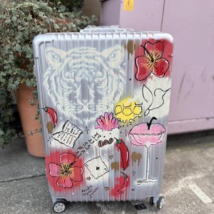 May include: A silver suitcase with a white tiger face painted on it. The suitcase also has red flowers, chilli peppers, a playing card, and the text "Live the Life You Love, Love the Life You Live" painted on it.
