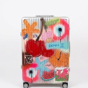 Könnte beinhalten: Ein silberner Hartschalenkoffer mit farbenfrohen Graffiti-Motiven, darunter rote Kirschen, ein orangefarbener Seestern, eine rosa Muschel, ein Stück Wassermelone, ein Koala, ein roter Drache sowie der Schriftzug Gemini II und HYROX in Sprühfarbe.