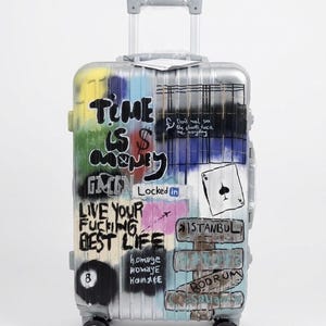 May include: A silver hard-shell carry-on suitcase with four wheels and a retractable handle. The exterior features colourful graffiti-style spray paint, including the text "TIME IS MONEY," "LIVE YOUR FUCKING BEST LIFE," "ISTANBUL," and "BODRUM."