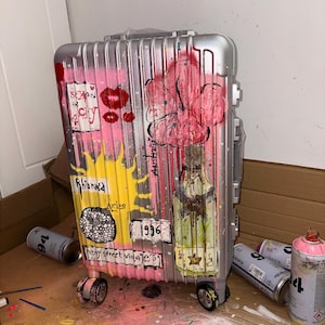 May include: A silver hard-shell suitcase featuring vertical ridges and custom graffiti-style artwork. The design includes pink and yellow paint splashes, a large pink flower, red lips, and text reading Sex and the City, Rihanna, Aries, 1996, and Australia.