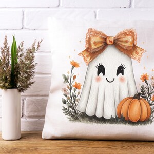May include: White tote bag featuring a cute ghost illustration with a bow, blush cheeks, and a smiling face. An orange pumpkin and flowers are at the bottom. A vase with decorative grass is on the left.