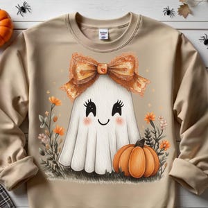 May include: Beige sweatshirt featuring a cute ghost with a bow, surrounded by flowers and a pumpkin. The ghost has a smiling face and is wearing a large orange bow. The design is perfect for Halloween.