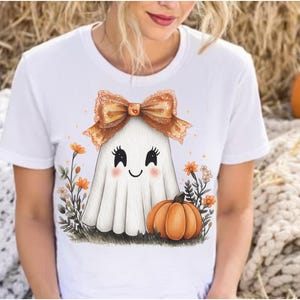 May include: White t-shirt featuring a cute ghost character with a bow, blush, and a pumpkin. The ghost is surrounded by flowers and grass, creating a festive Halloween design.