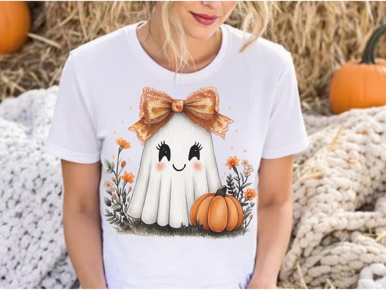May include: White t-shirt featuring a cute ghost character with a bow, blush, and a pumpkin. The ghost is surrounded by flowers and grass, creating a festive Halloween design.