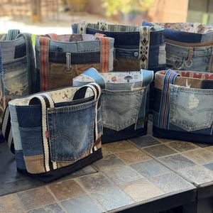 May include: A collection of tote bags crafted from repurposed blue denim jeans, featuring various shades of indigo, integrated pockets, fabric straps, and decorative woven trim accents. Each bag displays unique patchwork construction and visible stitching details.