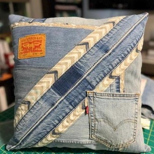 May include: A square throw pillow constructed from light blue denim fabric featuring a diagonal pattern of beige chevron ribbon. The pillow includes a tan Levi Strauss & Co. leather patch in the upper left corner and a denim pocket on the lower right.
