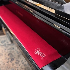 May include: A red piano key cover with a white musical note design. The cover is rectangular and fits inside the piano's key bed. The piano is black and shiny.