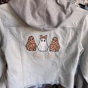 May include: Light blue denim jacket with a gray hood. The back features three embroidered ghost patches, two with leopard print and one white with a leopard print bow. The text "Boo-Jee" is embroidered below the ghosts.