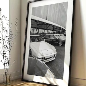 May include: Black and white framed print featuring classic cars parked in front of a modern building. The image showcases a white sports car in the foreground, with other vintage vehicles visible in the background. The photograph is displayed in a black frame.