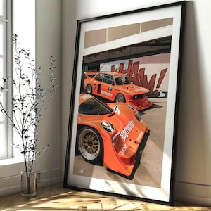 May include: Framed print featuring two orange race cars with sponsor logos. The cars are parked in front of a wall with graffiti-style lettering. The print is in a black frame and leans against a wall.