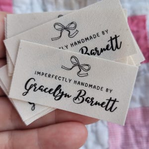 May include: Several off-white fabric labels with black text and a bow graphic. The labels read "Imperfectly Handmade by Gracelyn Barnett". The labels are stacked and held in a hand, with a colorful patchwork quilt in the background.