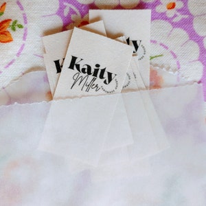 May include: A stack of white fabric tags with black text. The top tag reads "Kaity Miller Handmade Goods".