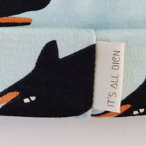 Puede incluir: Un gorro azul claro con un patr&oacute;n de ballena negra y el texto "IT'S ALL BIG'N" en una etiqueta blanca.