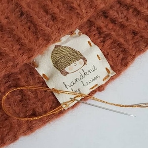 May include: A close-up of a brown knitted hat with a white label sewn on. The label features a cartoon illustration of a person wearing a hat and the text "handknit by Lauren".