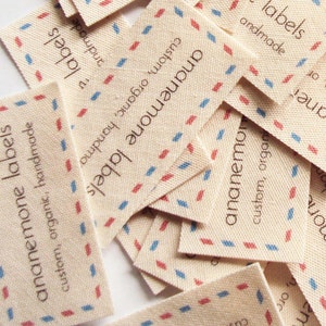 May include: A pile of white fabric labels with a blue and red diamond pattern border. The labels say "ananemone labels" and "custom, organic, handmade".