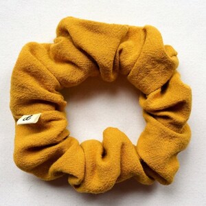 Puede incluir: Un scrunchie de color amarillo mostaza hecho de tela. El scrunchie tiene una peque&ntilde;a etiqueta blanca con la letra "d" en ella.