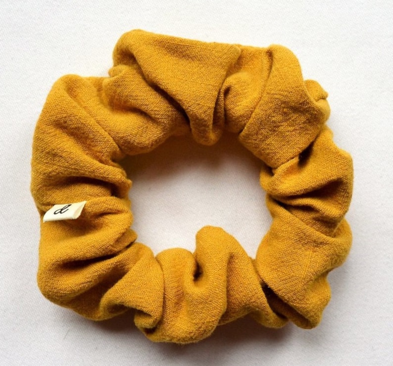 Puede incluir: Un scrunchie de color amarillo mostaza hecho de tela. El scrunchie tiene una peque&ntilde;a etiqueta blanca con la letra "d" en ella.