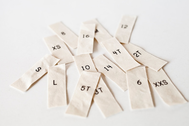 May include: A collection of white fabric tags with various sizes and numbers printed on them, including XS, S, L, 4T, 5T, 6, 10, 12, 14, 2T, and 3T.