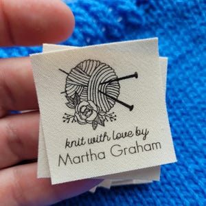 Puede incluir: Etiqueta de tela blanca con una ilustración en blanco y negro de un ovillo de lana con agujas de tejer y una flor. El texto "knit with love by Martha Graham" está impreso debajo de la ilustración.