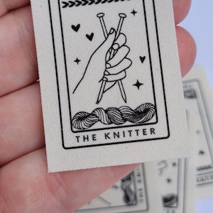 Pode incluir: Uma etiqueta de tecido branco com texto preto e um gr&aacute;fico. O texto diz "Lauren O'Shea" e "The Knitter". O gr&aacute;fico &eacute; uma m&atilde;o segurando duas agulhas de tric&ocirc; com fio.