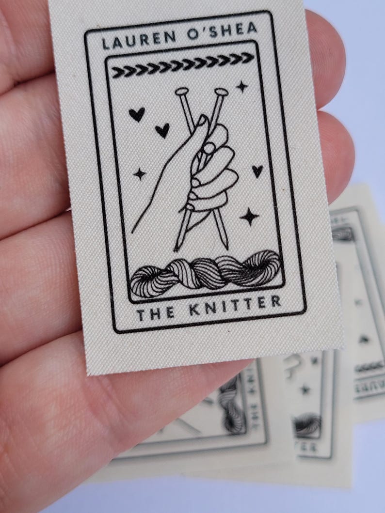 Pode incluir: Uma etiqueta de tecido branco com texto preto e um gr&aacute;fico. O texto diz "Lauren O'Shea" e "The Knitter". O gr&aacute;fico &eacute; uma m&atilde;o segurando duas agulhas de tric&ocirc; com fio.