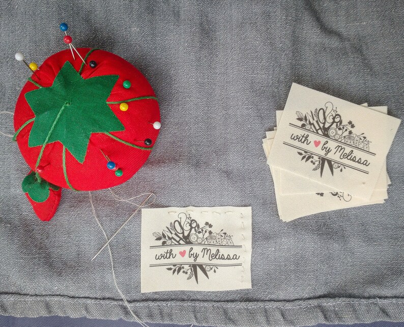 May include: A red tomato-shaped pin cushion with a green leaf design, holding several colorful pins. Beside it are several fabric labels with the text "with by Melissa" and a floral and scissors design, perfect for sewing projects.