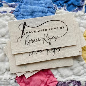 Puede incluir: Un montón de etiquetas de tela blanquecina con texto negro. Las etiquetas dicen "MADE WITH LOVE BY Grace Keyes" con un gráfico de aguja e hilo. Las etiquetas están sobre un edredón de retazos colorido.