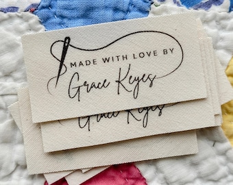 Peut inclure: Plusieurs étiquettes en tissu blanc cassé avec du texte noir. Elles indiquent "MADE WITH LOVE BY Grace Keyes" avec un motif d'aiguille et de fil. Les étiquettes sont posées sur une courtepointe colorée.
