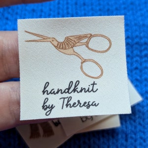 Puede incluir: Una etiqueta de tela blanca con una ilustraci&oacute;n marr&oacute;n de un par de tijeras. El texto "handknit by Theresa" est&aacute; impreso en negro debajo de la ilustraci&oacute;n.
