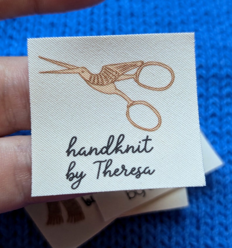 Puede incluir: Una etiqueta de tela blanca con una ilustraci&oacute;n marr&oacute;n de un par de tijeras. El texto "handknit by Theresa" est&aacute; impreso en negro debajo de la ilustraci&oacute;n.