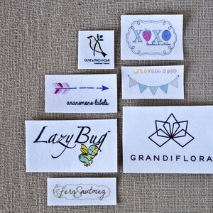 May include: A collection of six different logo designs printed on white fabric labels. The logos include a bird, an arrow, a butterfly, a flower, a heart, and a string of flags. The text on the labels includes "Olive & Finch Home", "ananemare labels", "Lazy Bug", "GRANDIFLORA", "XOXO handmade with love", and "Little Peek a boo".
