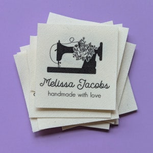 Peut inclure: Une pile d'&eacute;tiquettes en tissu blanc avec de l'encre noire. L'&eacute;tiquette repr&eacute;sente une silhouette de machine &agrave; coudre avec un motif floral et le texte "Melissa Jacobs handmade with love".