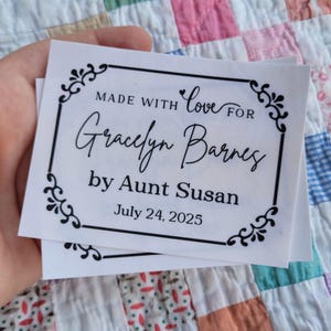 Puede incluir: Dos etiquetas rectangulares blancas con bordes decorativos negros y texto. El texto dice "MADE WITH love FOR Gracelyn Barnes by Aunt Susan July 24, 2025". Las etiquetas se sostienen en una mano.