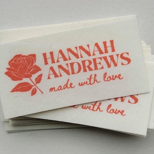 以下が含まれることがあります： 赤いバラと「HANNAH ANDREWS made with love」という文字が印刷された白い布製のタグが積み重ねられています。