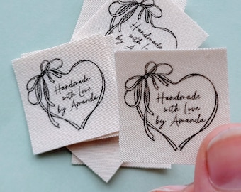 May include: Off-white fabric labels with a black heart-shaped design and a bow. The text "Handmade with Love by Amanda" is printed inside the heart. Several labels are visible, some stacked, with one held by a hand.