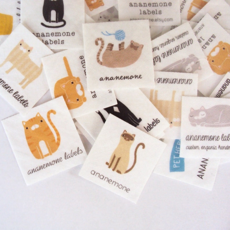 Puede incluir: Un conjunto de etiquetas de tela blanca con ilustraciones de gatos en naranja, marr&oacute;n, negro y gris. Cada etiqueta presenta la palabra "ananemone", y algunas incluyen la palabra "labels". Las etiquetas est&aacute;n dispersas sobre una superficie blanca.