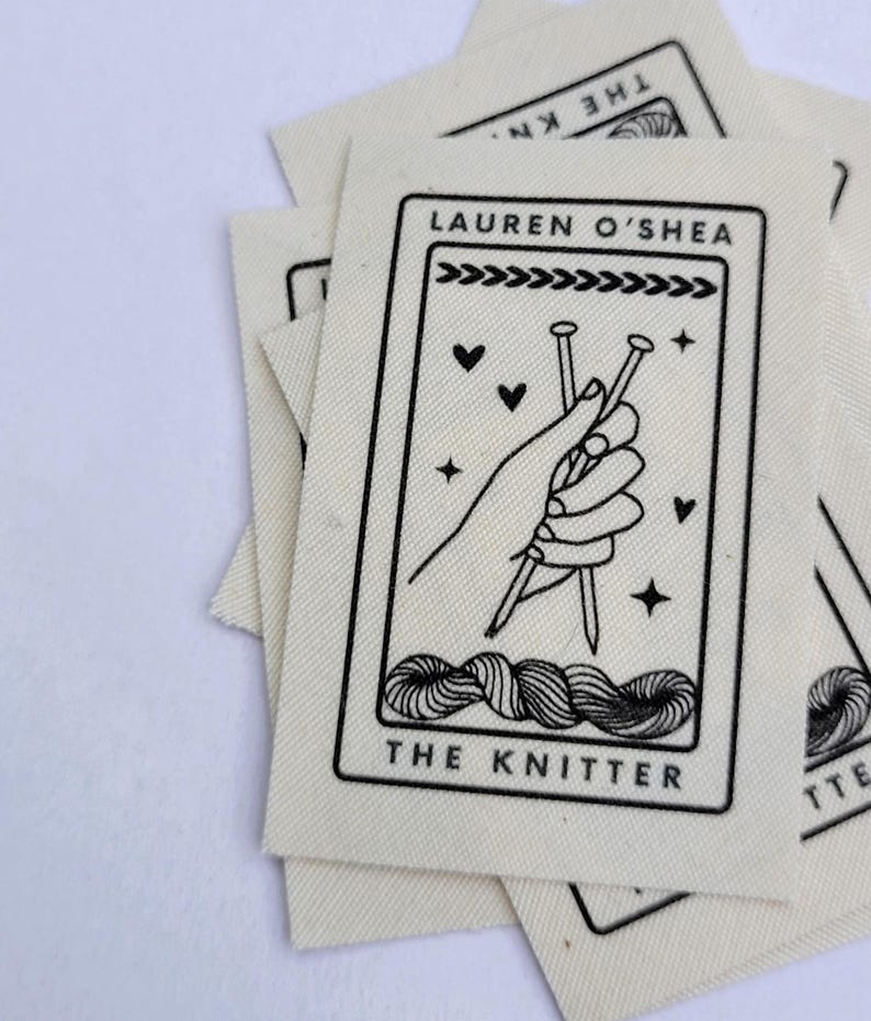 Pode incluir: Um remendo de tecido branco com texto preto e um gr&aacute;fico de uma m&atilde;o segurando agulhas de tric&ocirc;. O texto diz "Lauren O'Shea The Knitter".
