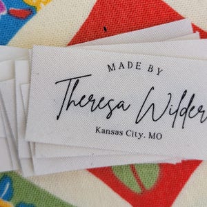 Può includere: Un mucchio di etichette in tessuto bianco con testo nero che recita "MADE BY Theresa Wilder Kansas City, MO".
