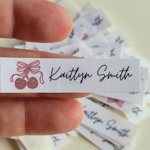May include: White fabric labels with the name "Kaitlyn Smith" in black script. Each label features a pink bow and cherry design. A hand holds one label, with more labels in the background.